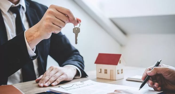 Man in suit hold key out over desk with house on it