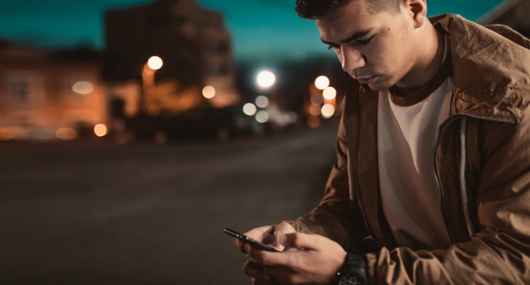 A teenager looking at their smart phone on a street at night time.